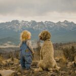 A child and dog sit together facing a vast mountain range, enjoying a serene moment.