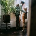 A man and an Akita dog share a tender moment by the window in a cozy apartment filled with plants.